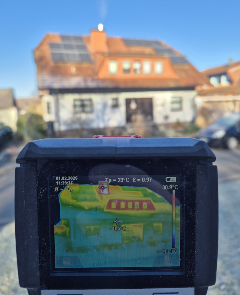 Wärmebildkamera im Vordergrund, im Display die Thermografie eines Wohnhauses mit Solaranlage